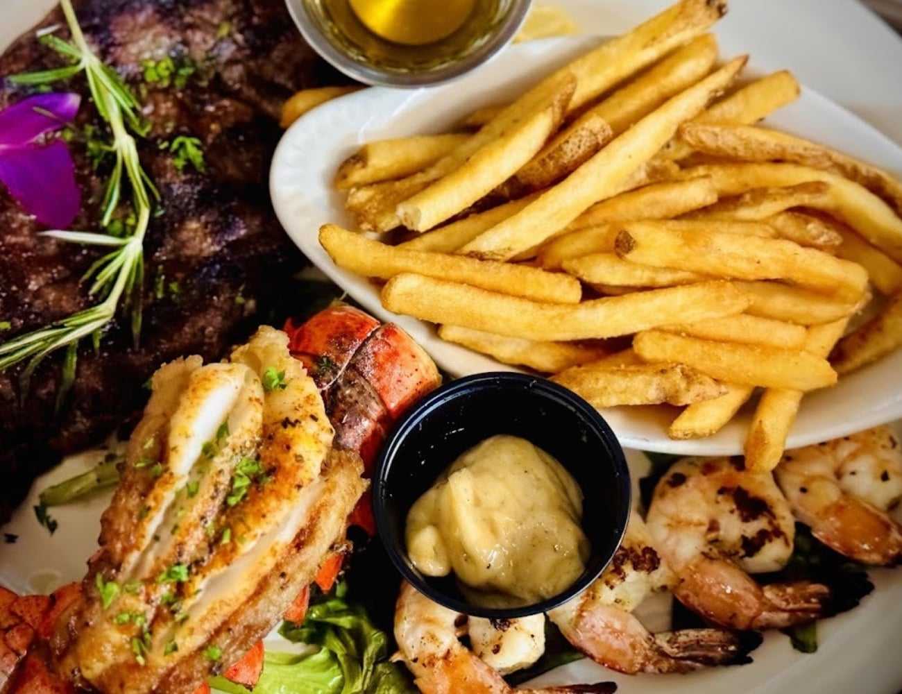 Steak with fries, grilled shrimp, lobster tail, and aioli on a white plate.